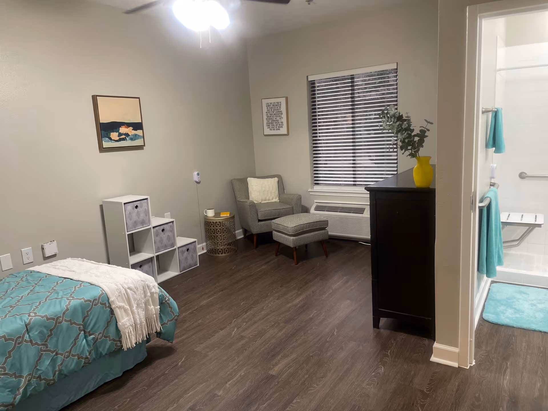A cozy assisted living bedroom with a bed covered in a teal and white patterned blanket and a white throw. There is a small white storage unit with fabric bins, a round side table with a cup and book, and a gray armchair with an ottoman near a window with blinds. A dark wooden dresser with a yellow vase and greenery is next to an open doorway leading to a bathroom with a shower, grab bars, and teal towels and bath mat. The room has wood flooring and neutral-colored walls with two framed pictures.