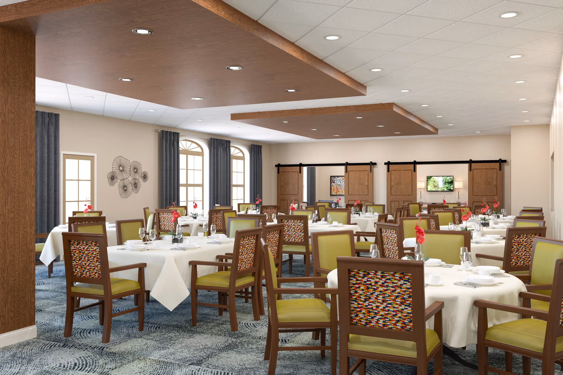 A spacious dining room with multiple round tables covered in white tablecloths, each set with plates, glasses, and red flower centerpieces. The chairs have wooden frames with green cushions and colorful patterned backs. The room features large windows with dark curtains, decorative wall art, and wooden sliding barn doors on one wall. The ceiling has recessed lighting and wooden accent panels.