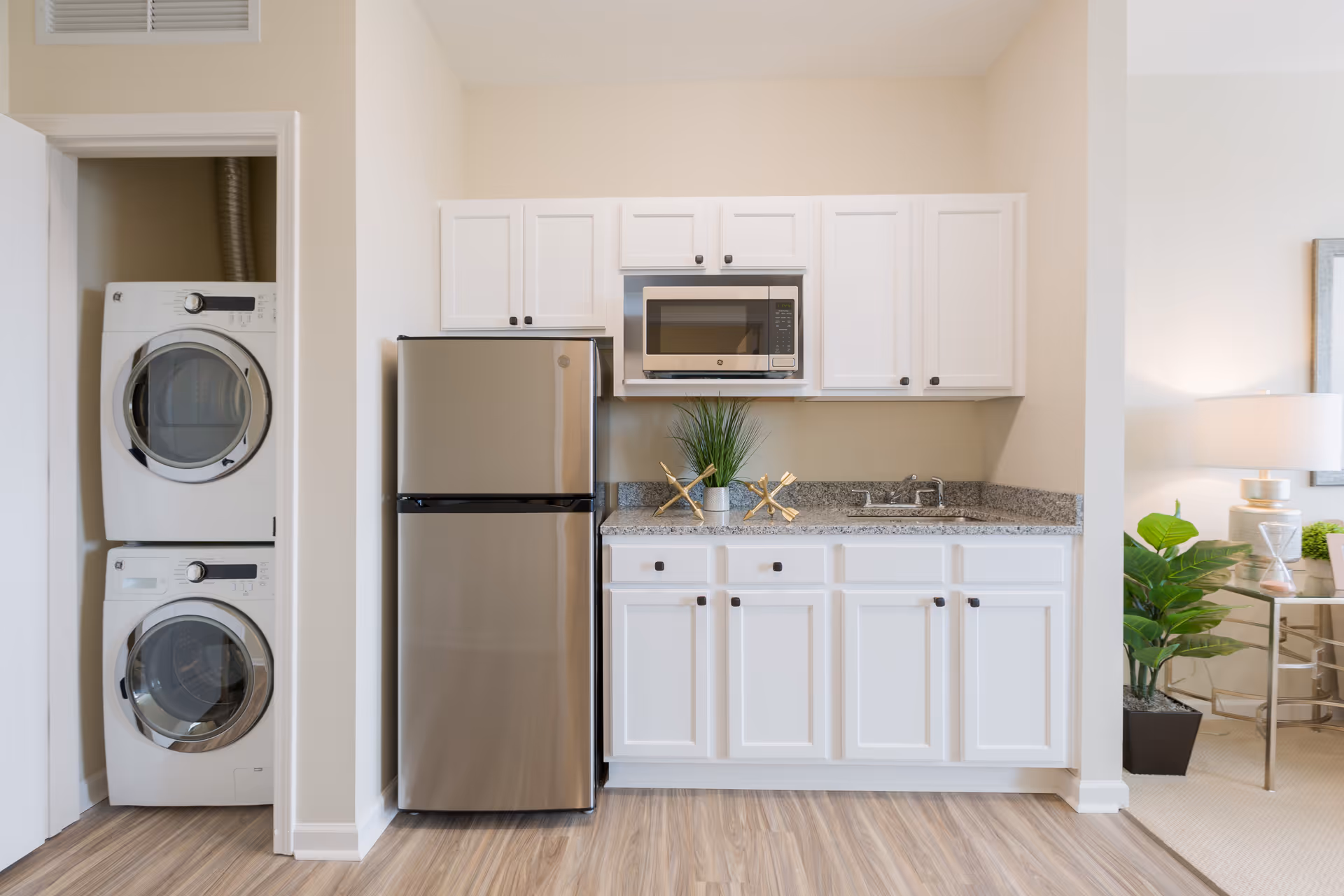 A compact kitchen area with white cabinets, a stainless steel refrigerator, a microwave mounted above the counter, and a sink. To the left, there is a small laundry closet with a stacked washer and dryer. On the right side, part of a living room area is visible with a lamp, a plant, and a side table.