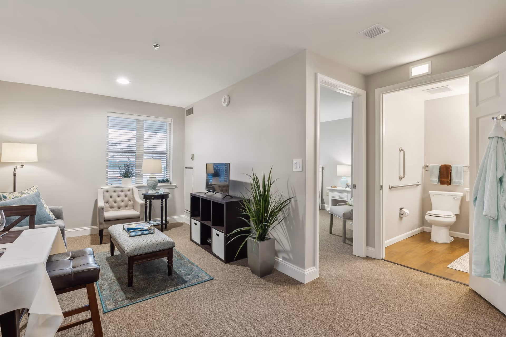A cozy living room area with a beige armchair, a small round table with a lamp and plant, a cushioned ottoman on a blue rug, a TV on a dark wooden shelf, and a tall green plant in a gray pot. To the right, an open door reveals a bathroom with a toilet, grab bars, towels, and a bathrobe hanging on the door. Another doorway leads to a bedroom with a nightstand and lamp visible.