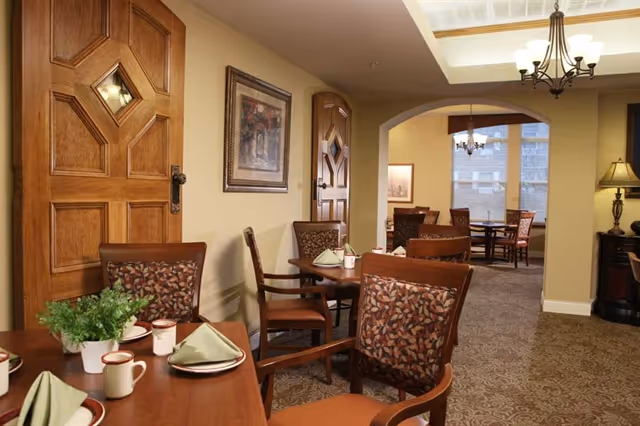 Interior view of a dining area in a senior living facility with wooden tables and chairs, each table set with green napkins, cups, and plates. The room features warm beige walls, decorative wooden doors, framed artwork, carpeted floors, and ceiling light fixtures including a chandelier and a lamp on a side table.
