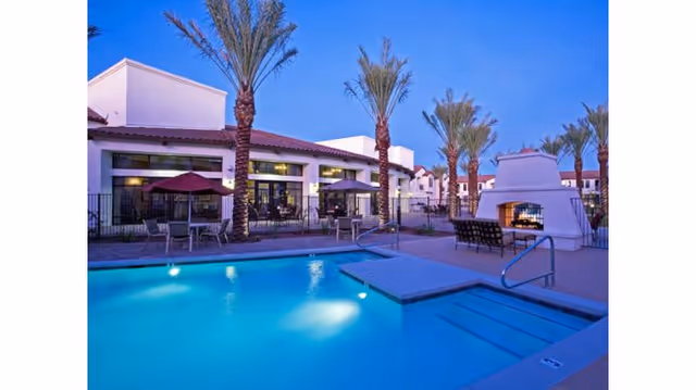 Outdoor pool area at dusk with palm trees, patio tables with umbrellas, and a white outdoor fireplace. The building in the background has large windows and a tiled roof.