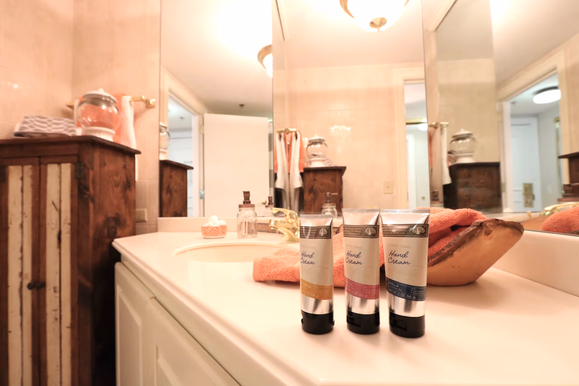 Bathroom countertop with three tubes of hand cream, a soap dispenser, a tissue box, and a wooden decorative bowl with an orange towel. A wooden cabinet and a large mirror are visible in the background, reflecting the open door and hallway.