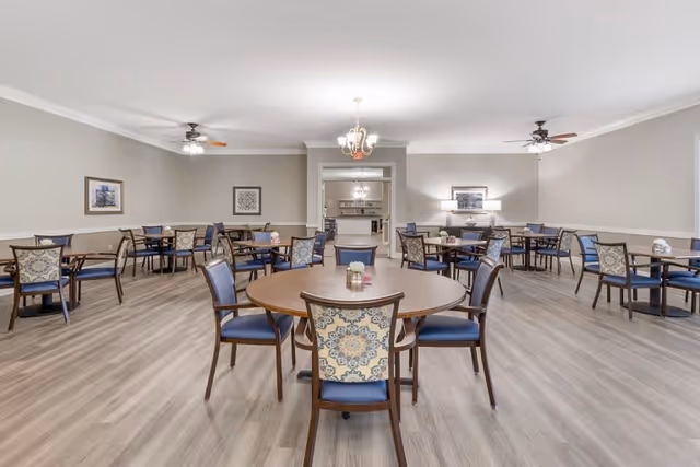 A spacious senior living dining room with multiple round and square wooden tables surrounded by chairs with blue cushions and patterned backs. The room has light-colored walls, wood flooring, ceiling fans with lights, and framed artwork on the walls. A chandelier hangs from the ceiling in the center, and there is a doorway leading to another room in the background.
