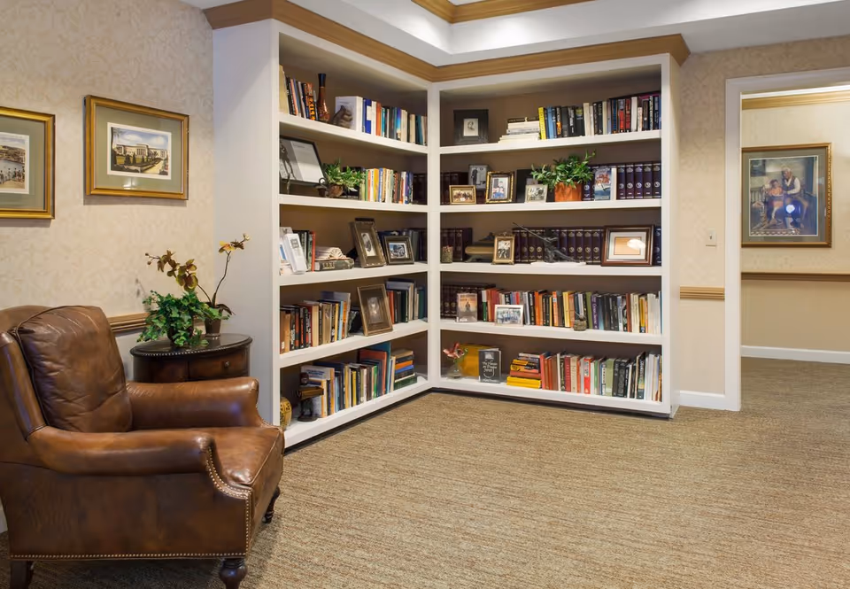 Cozy corner of a living room or common area with a brown leather armchair next to a small round wooden table holding a potted plant. Behind the chair is a built-in white bookshelf filled with books, framed photos, and decorative plants. The walls are decorated with framed artwork and have a beige patterned wallpaper. The floor is carpeted in a neutral tone.