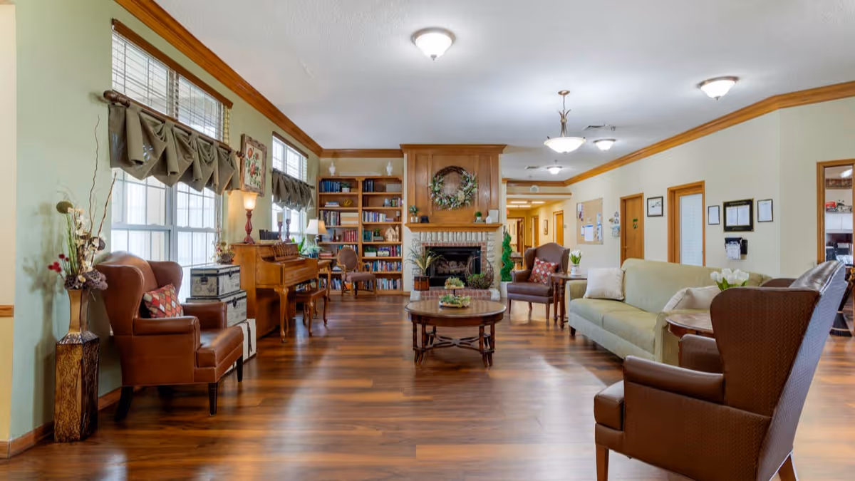 Spacious, well-lit senior living common room with seating, a fireplace, bookshelves, a piano, and hardwood floors.