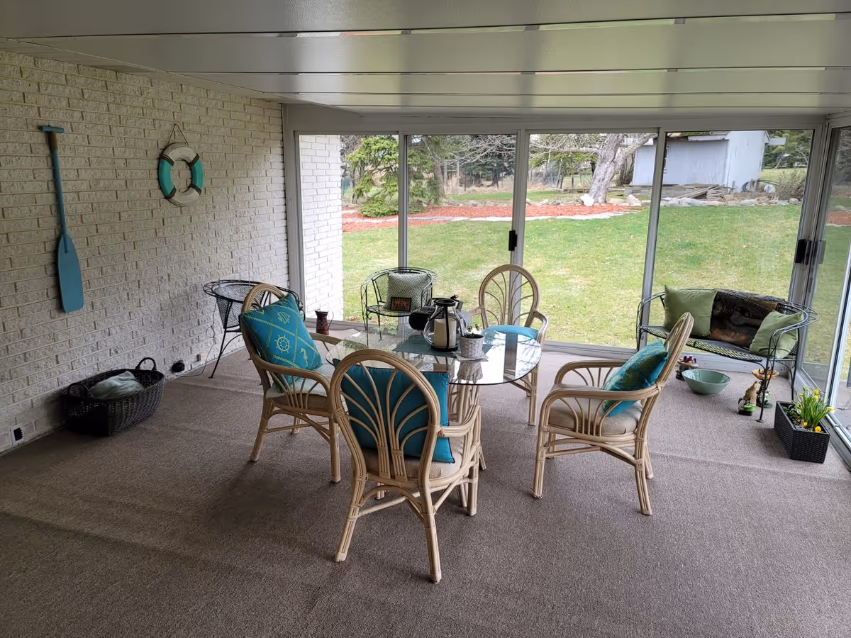 A sunroom with a glass table surrounded by four wicker chairs with blue and green cushions. The room has large sliding glass doors that open to a grassy backyard with trees and a small shed. The walls are white brick, decorated with a blue paddle and a life preserver. There are additional chairs and decorative items along the edges of the room.