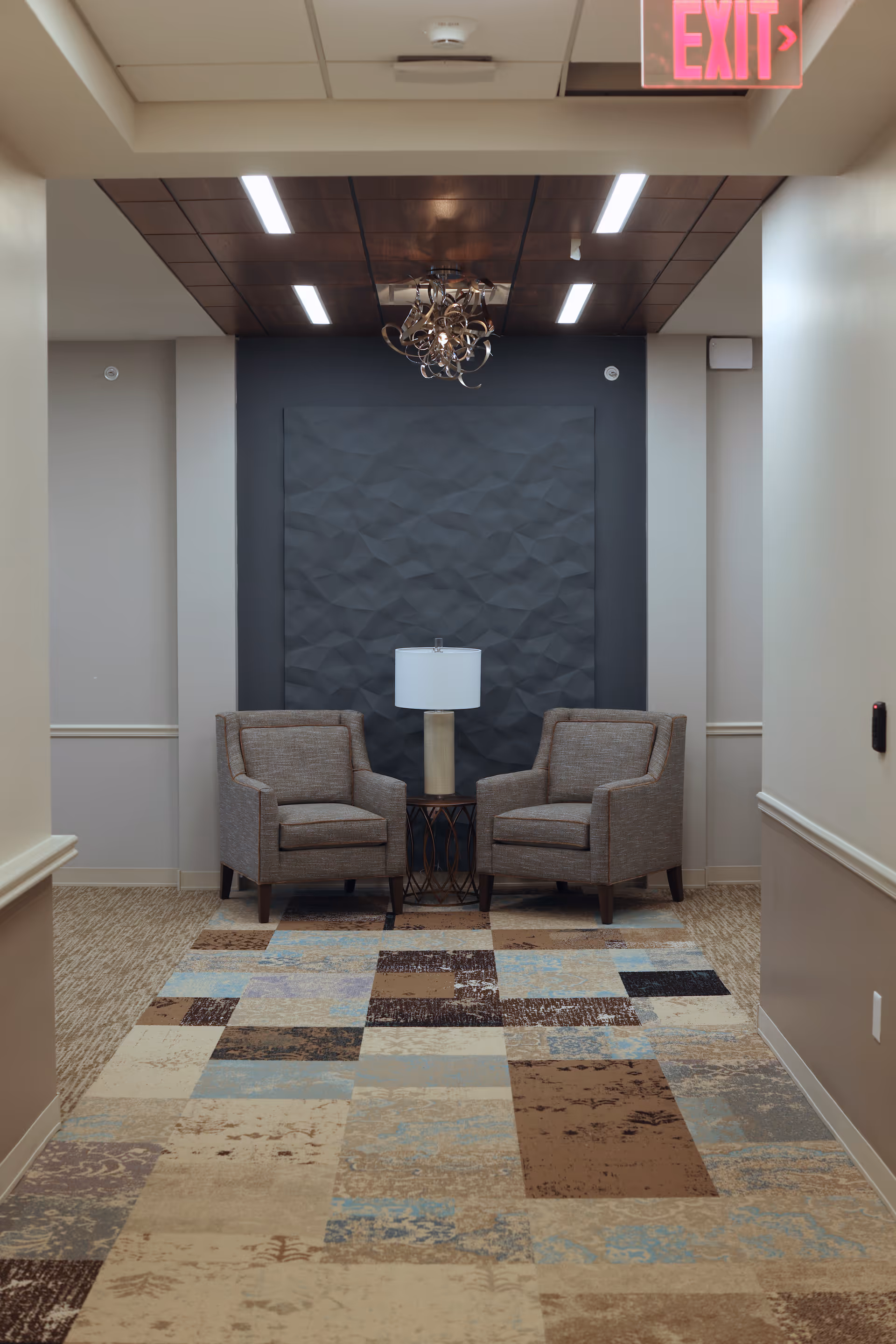A seating area in a hallway with two gray upholstered armchairs separated by a small round table with a lamp. The wall behind features a textured dark panel, and the ceiling has recessed lighting and a decorative chandelier. The floor is covered with a patterned carpet in shades of beige, brown, blue, and cream. An illuminated exit sign is visible at the top right corner.