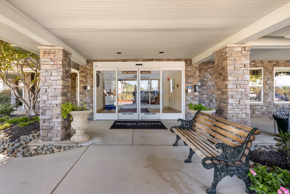 Covered entrance to Shasta Estates Senior Living with stone pillars, a wooden bench with decorative metal armrests, potted plants, and glass automatic doors leading inside.