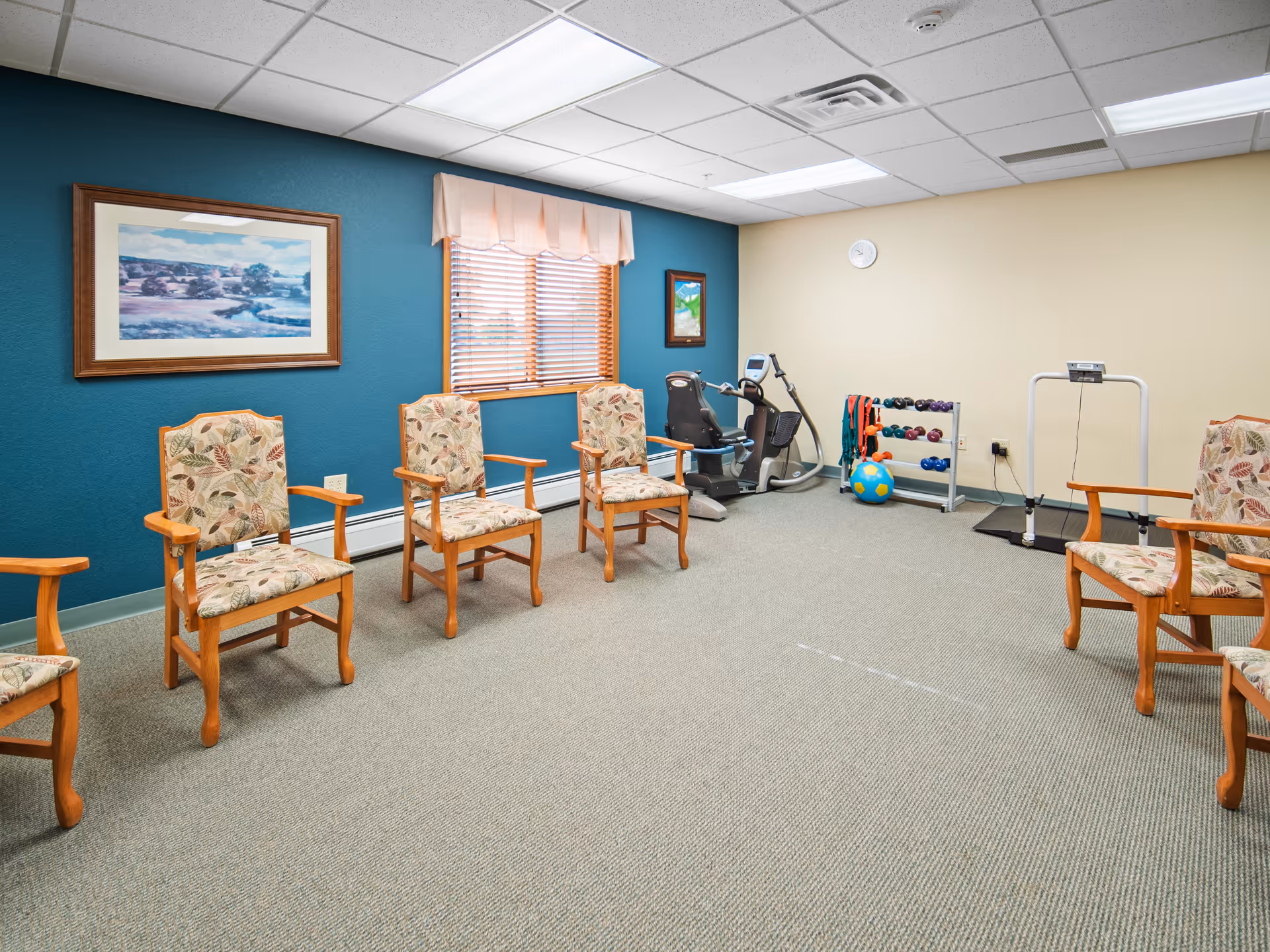A senior living activity room with wooden armchairs arranged around the perimeter and exercise equipment and weights against teal and beige walls.