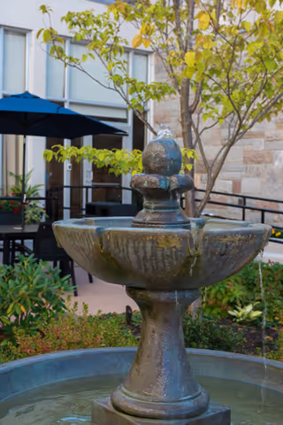 A close-up view of a stone water fountain in an outdoor garden area with green plants, a tree, and patio furniture including a table and chairs under a blue umbrella. The background shows part of a building with windows and stone walls.