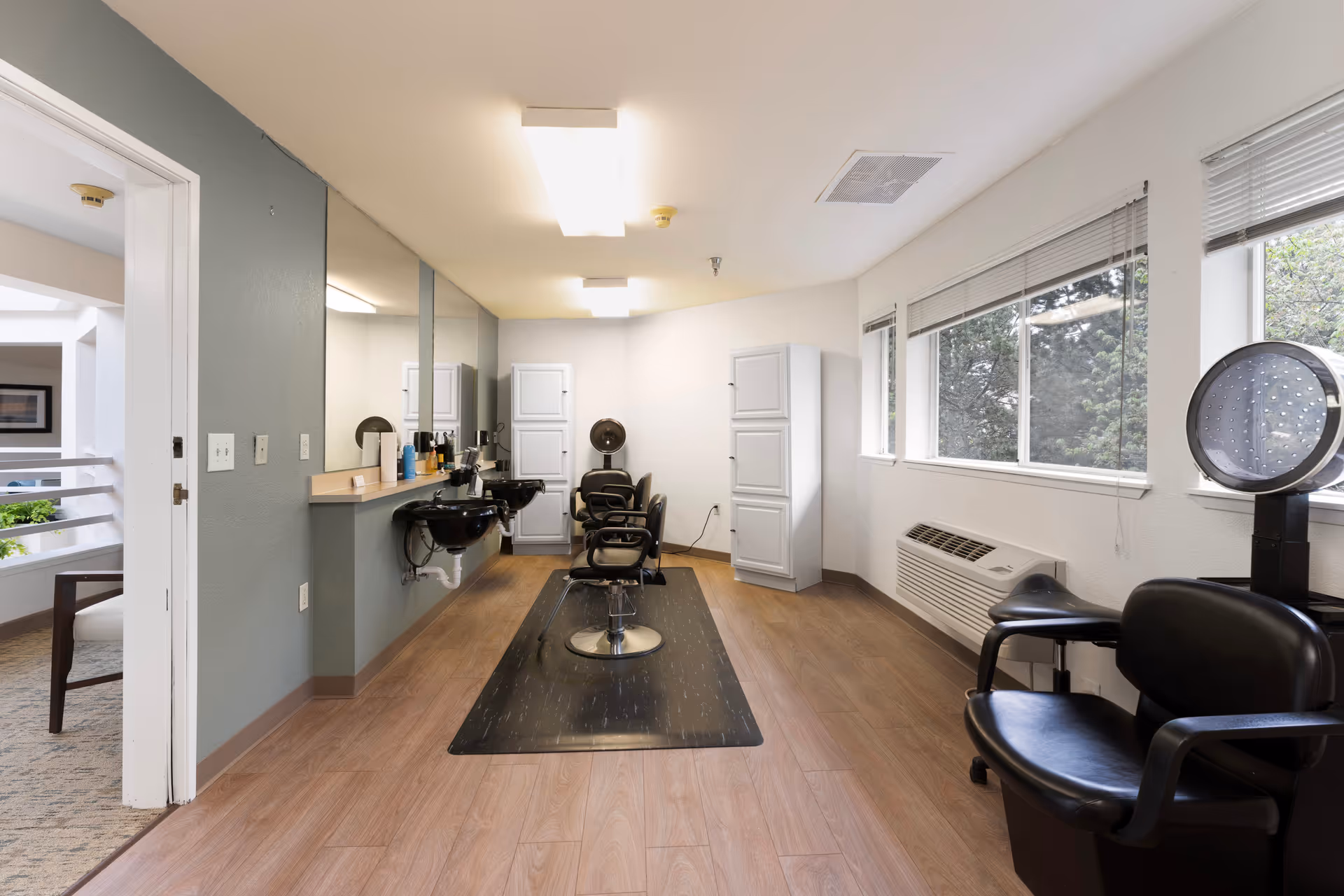 Interior view of a hair salon area in a senior living facility with two black salon chairs in front of a long mirror and two black sinks. There are white cabinets at the back wall, a hair dryer chair on the right side near windows with blinds, and wood flooring throughout the room.