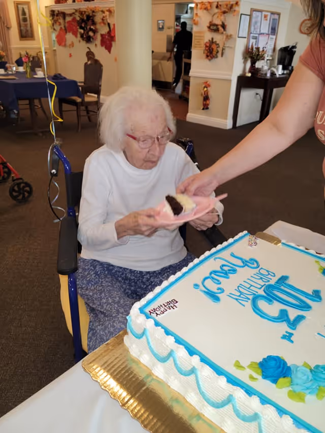 An elderly woman with white hair and glasses sitting in a wheelchair, holding a plate with a piece of cake being served to her by another person. In front of her is a large birthday cake with blue and white icing that reads 'Happy 103rd Birthday'. The setting appears to be a decorated common area with autumn-themed decorations on the walls.