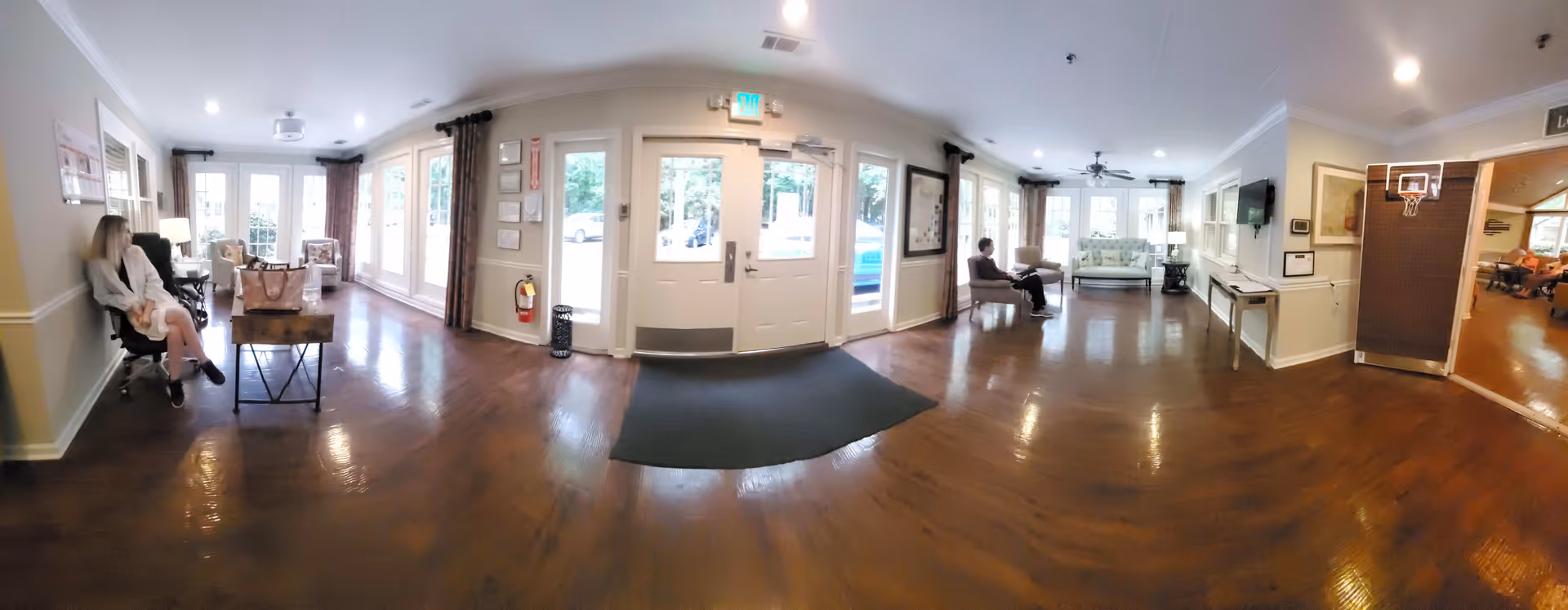 Wide view of a senior living facility common area with wooden floors, multiple seating areas including armchairs and a loveseat, large windows with curtains, and a glass door entrance. Two people are seated on opposite sides of the room. There is a basketball hoop mounted on a divider near an adjoining room where more people are visible.