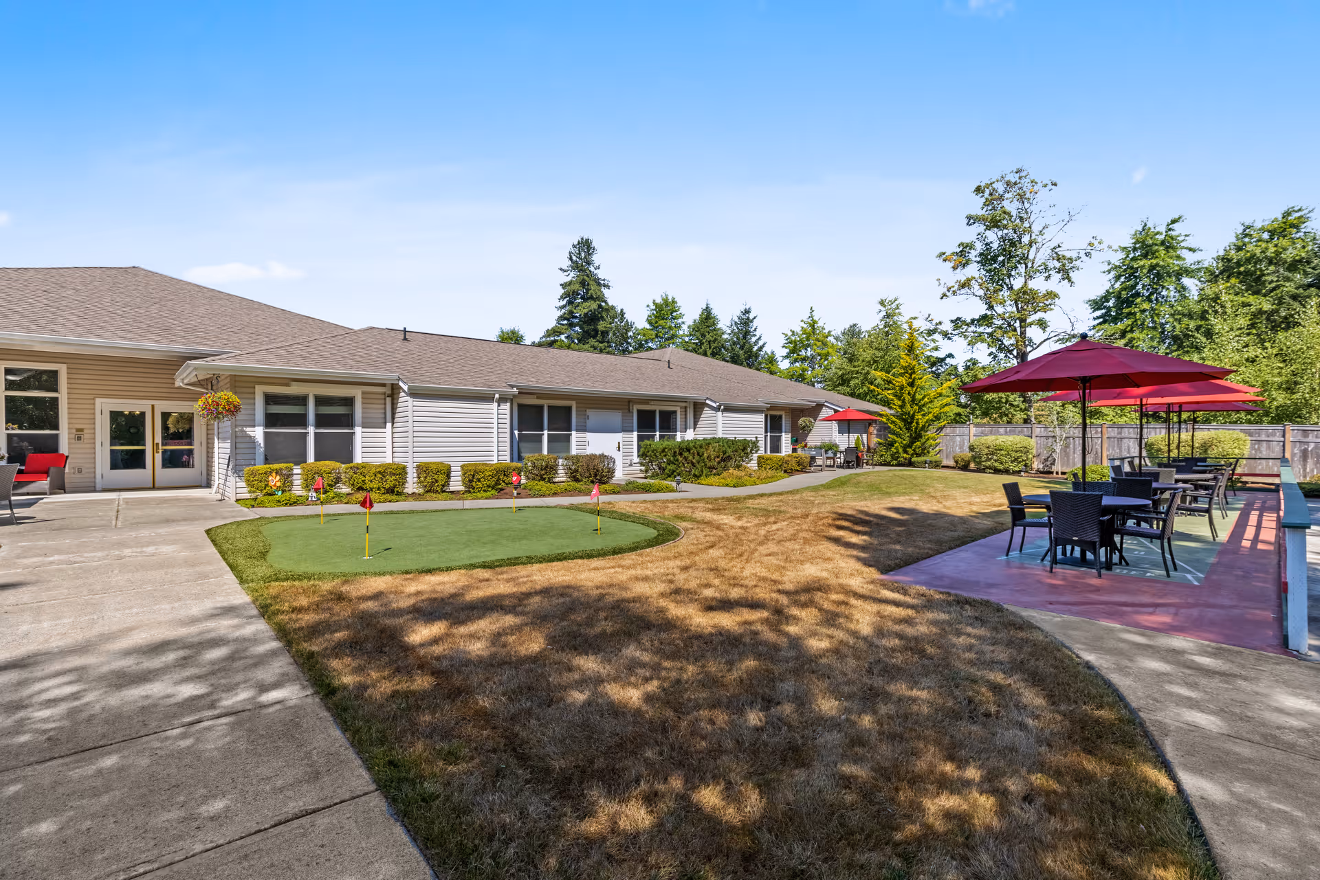 Outdoor area of a senior living facility with a small putting green, patio tables with red umbrellas, and a building with beige siding and multiple windows in the background under a clear blue sky.