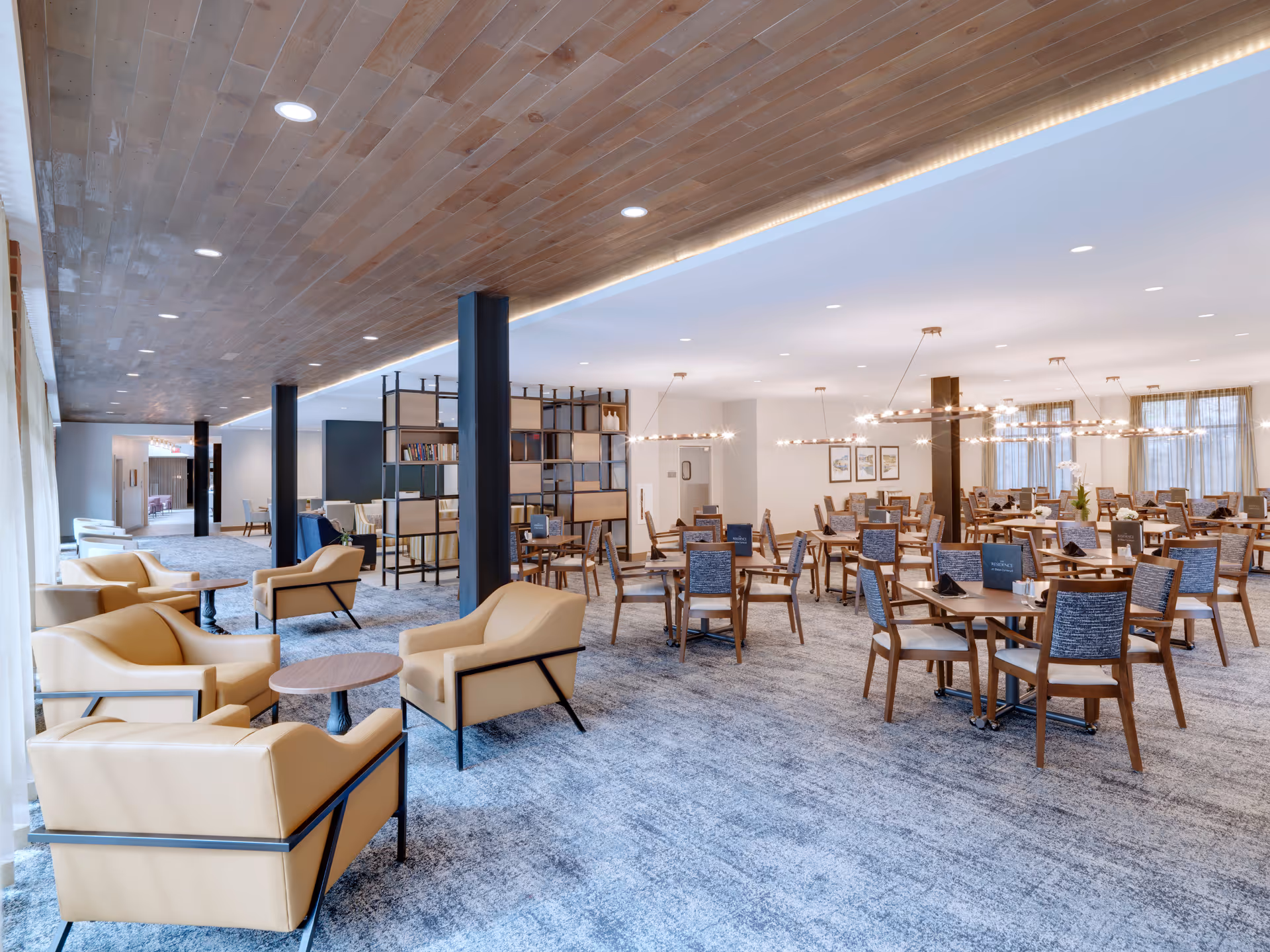 A spacious, modern dining area with multiple wooden tables and chairs arranged neatly. The room features beige armchairs with small round tables on the left side, a carpeted floor, and a wooden ceiling with recessed lighting. Large windows with sheer curtains allow natural light to fill the space. Decorative light fixtures hang from the ceiling, and a black metal shelving unit partially divides the seating areas.