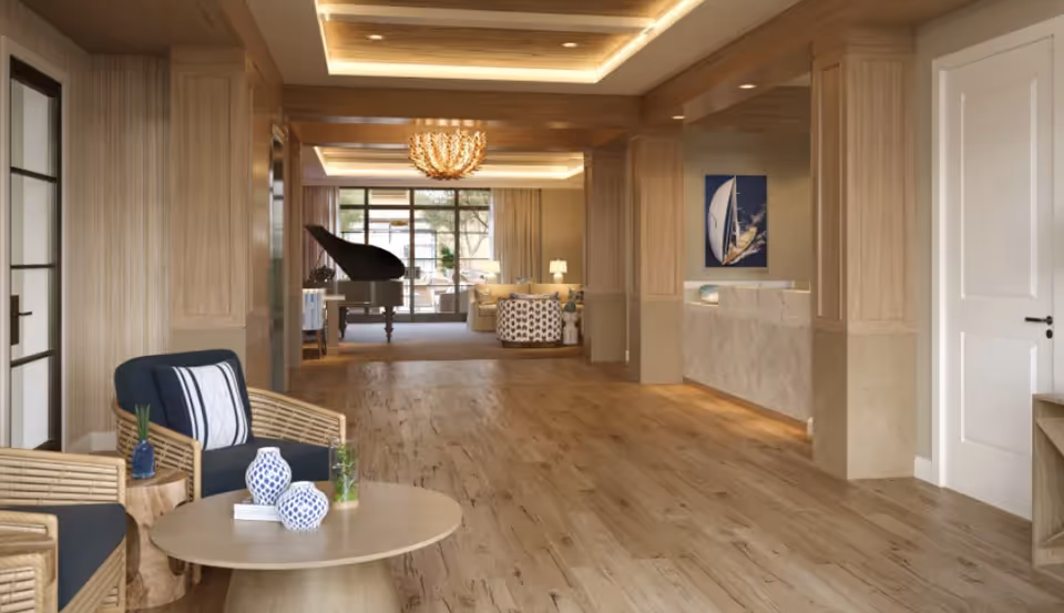 A spacious and elegant interior common area with wooden flooring and light wood-paneled walls. The room features comfortable seating with wicker chairs and navy cushions around a round wooden table with decorative vases. In the background, there is a grand piano, a chandelier, and a reception desk with a sailboat painting on the wall. Large windows allow natural light to fill the space.