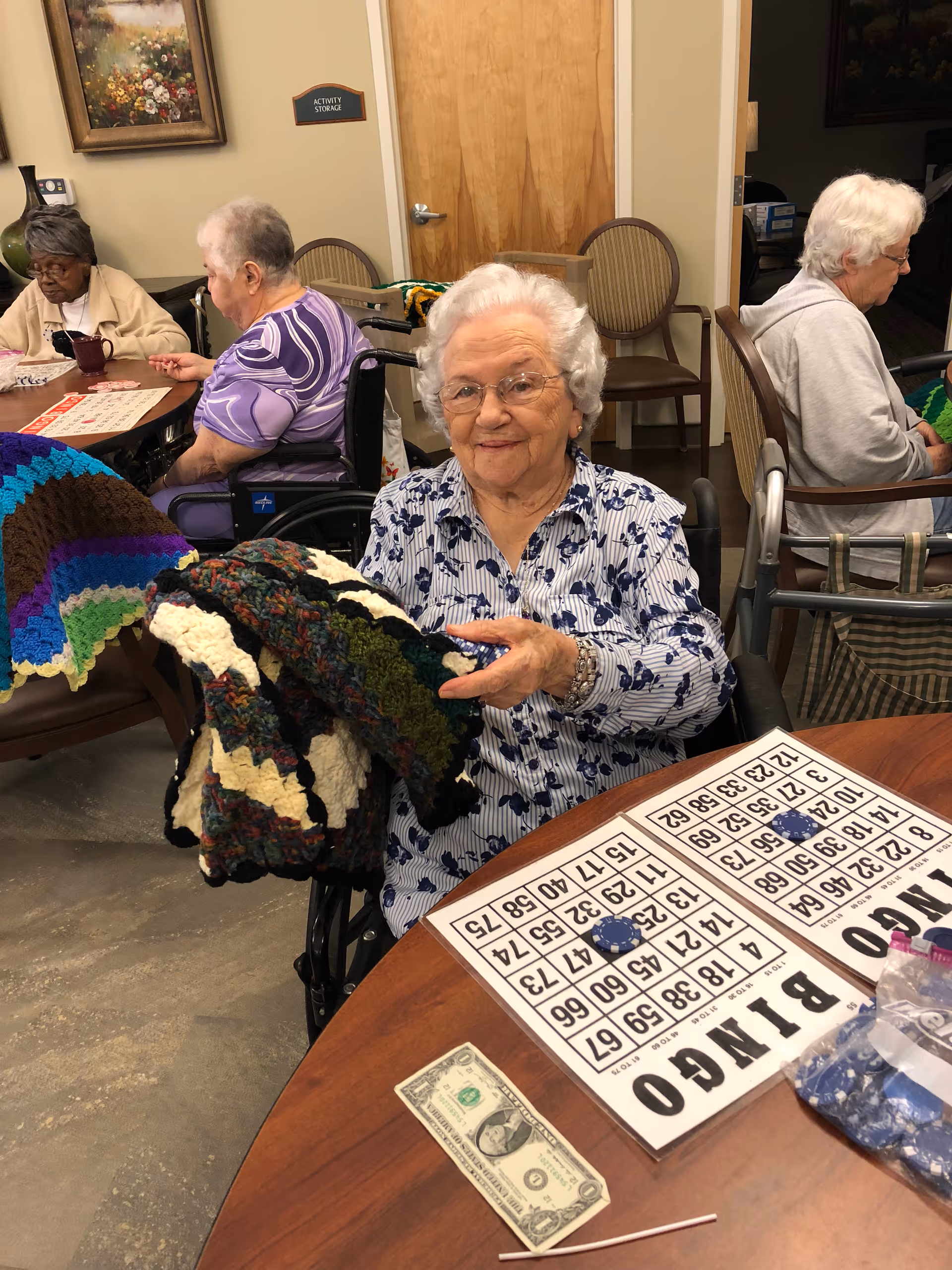 Elderly women sitting around a table in a senior living facility, playing bingo. One woman in the foreground is smiling and holding a colorful crocheted blanket. Bingo cards, chips, and a dollar bill are on the table. The room has beige walls, wooden doors, and chairs.