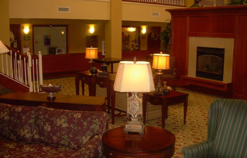 A cozy living room area in a senior living facility featuring patterned upholstered sofas and armchairs, wooden tables with decorative lamps, a fireplace with a wooden mantel, and warm lighting. The room has a carpeted floor with a floral pattern and a staircase with white railings in the background.