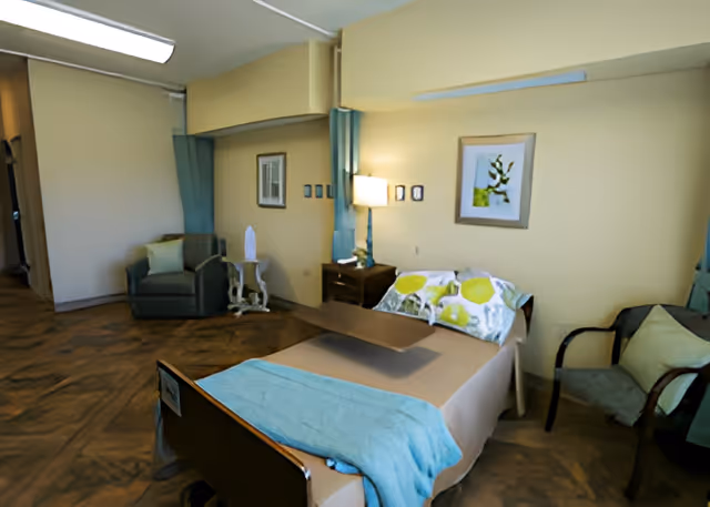 A furnished senior living bedroom with an adjustable bed, bedside table and lamp, multiple chairs, and wall art.