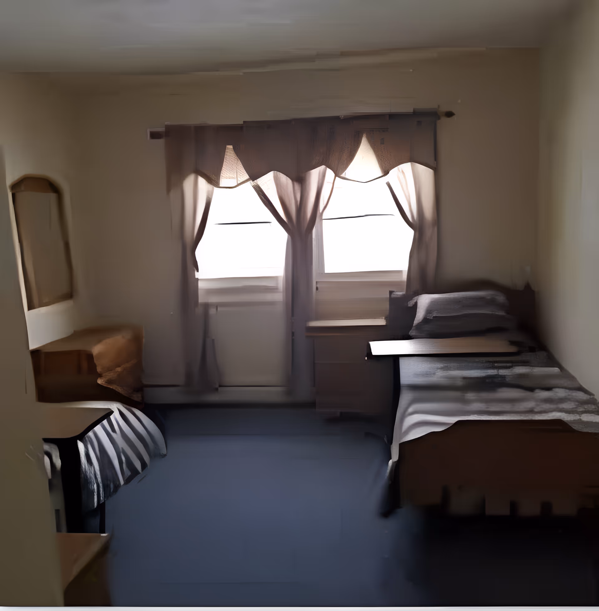 A small bedroom with two single beds on opposite sides of the room, each with a blanket and pillow. A window with sheer curtains is centered on the far wall, allowing natural light to enter. There is a small wooden desk and chair on the left side near one of the beds. The walls are painted a light color and the floor is covered with a dark carpet.