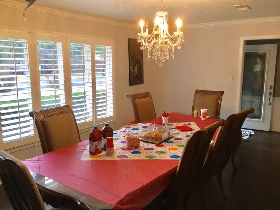 Dining room with a long table covered in a colorful party tablecloth and drinks, surrounded by upholstered chairs beneath a crystal chandelier.