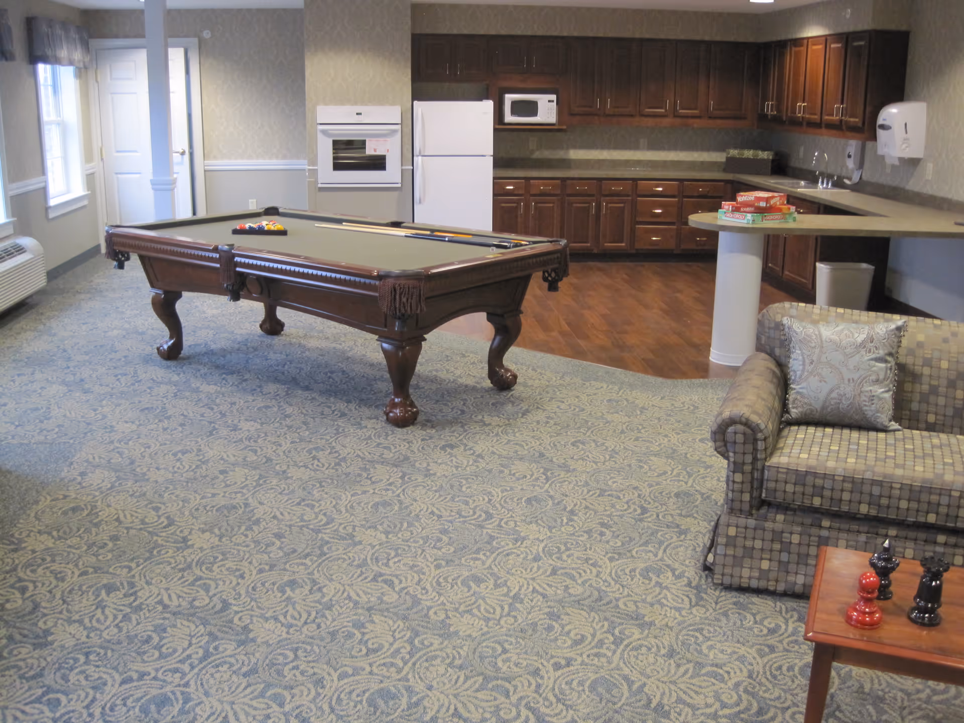A spacious room featuring a pool table with billiard balls and cues, a patterned carpet, a small seating area with a cushioned armchair and a wooden table with decorative chess pieces. The background shows a kitchenette with dark wooden cabinets, a white refrigerator, microwave, oven, and a counter with board games on it.