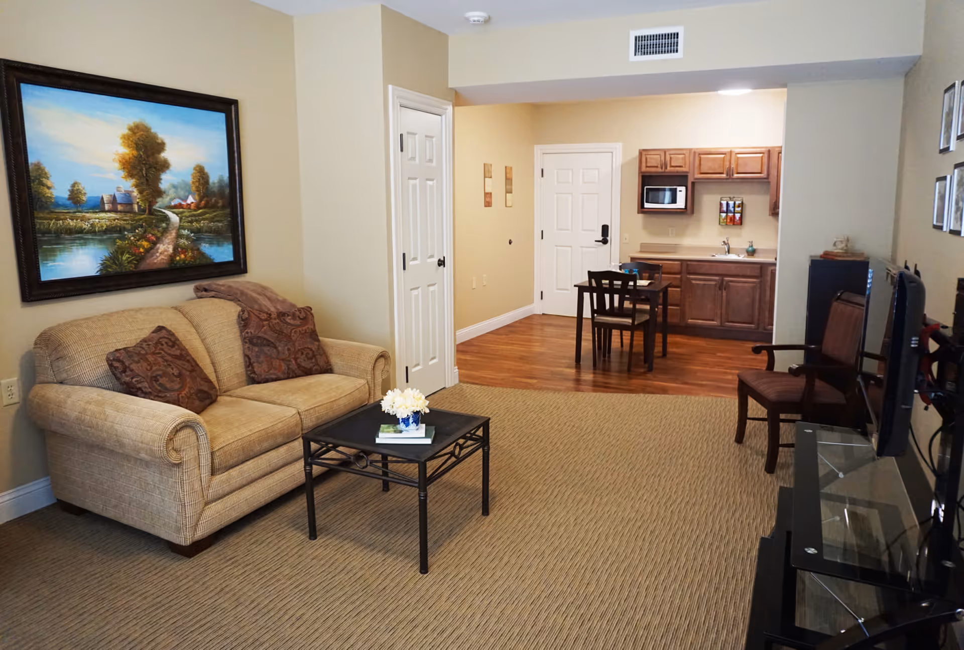 A cozy living area in an assisted living facility featuring a beige couch with two patterned pillows and a throw blanket, a small black coffee table with a flower vase and books, a wall painting of a scenic landscape, a dining table with two chairs, and a kitchenette with wooden cabinets, a microwave, and a sink. There is also a TV on a stand and a wooden chair near it.