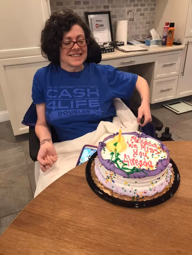 A smiling woman in a wheelchair wearing glasses and a blue New Jersey Lottery Cash 4 Life t-shirt is sitting at a table with a decorated cake in front of her. The cake has purple and white frosting with colorful sprinkles and a message written in red icing that says, 'Mekeda We Miss You Already.' The background shows a kitchen area with white cabinets and various items on the countertop.