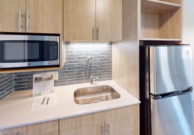 Modern kitchenette with a stainless steel refrigerator and microwave, a single-basin sink set in a white countertop, light wood cabinets, and a blue-gray tiled backsplash.