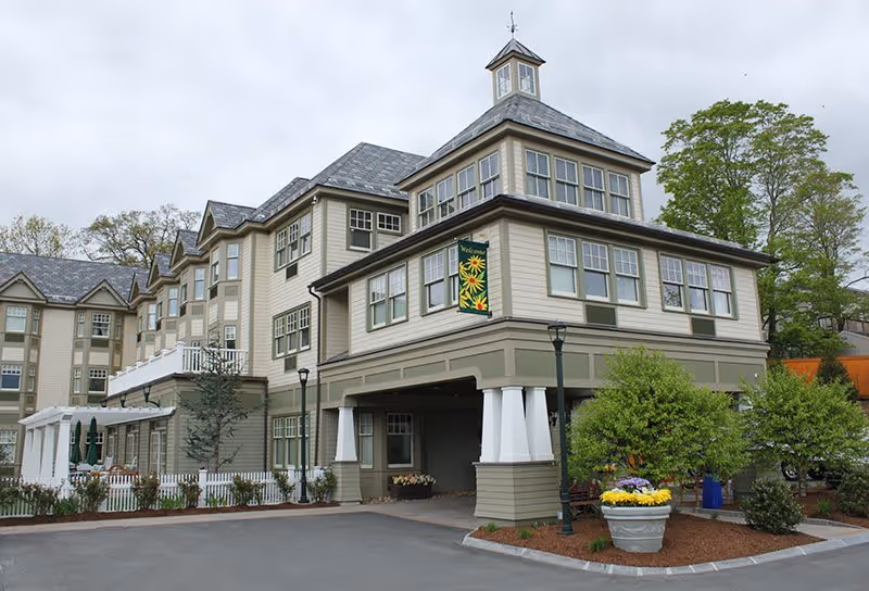 Exterior view of a multi-story assisted living facility building with beige siding and numerous windows. The building features a covered entrance supported by white columns, a small tower-like structure on the roof, and a decorative welcome banner with sunflowers. There are landscaped areas with trees, shrubs, and flower pots near the entrance, and a paved driveway in front.