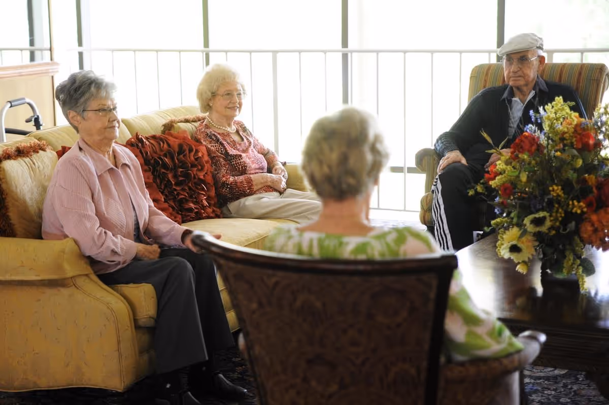 Four elderly individuals sitting and conversing in a well-lit living room area with large windows, a yellow couch, an armchair, and a coffee table with a colorful flower arrangement.