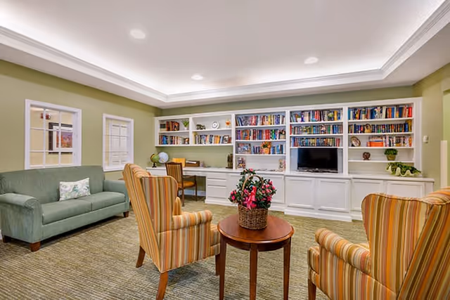 A cozy living room area in Brookdale Camino del Sol featuring a green sofa, two striped armchairs around a small round wooden table with a flower basket, built-in white bookshelves filled with books and decorative items, a small TV, and a desk with a chair. The walls are painted light green and the ceiling has recessed lighting.