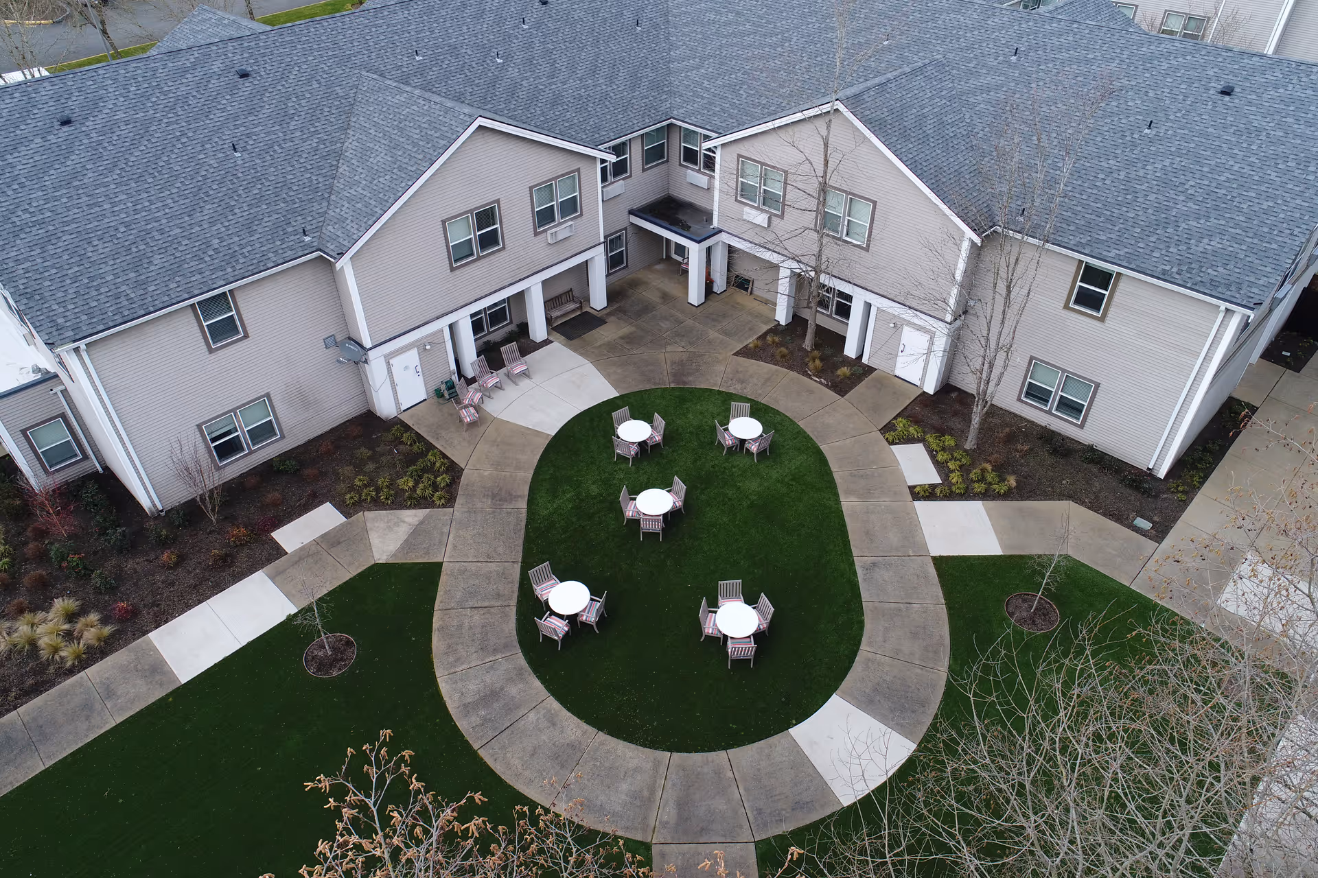 Aerial view of an outdoor courtyard area at GenCare Lifestyle Federal Way featuring a circular pathway surrounding a grassy area with five round tables, each accompanied by four chairs. The courtyard is adjacent to a two-story building with beige siding and multiple windows.