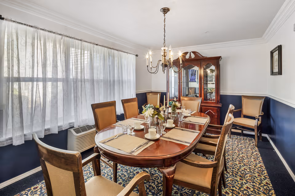 A formal dining room with a polished wooden table set for six people with placemats, cups, glasses, and a floral centerpiece. The room features a chandelier overhead, a large window with sheer white curtains, a patterned carpet, and a wooden china cabinet against the wall.