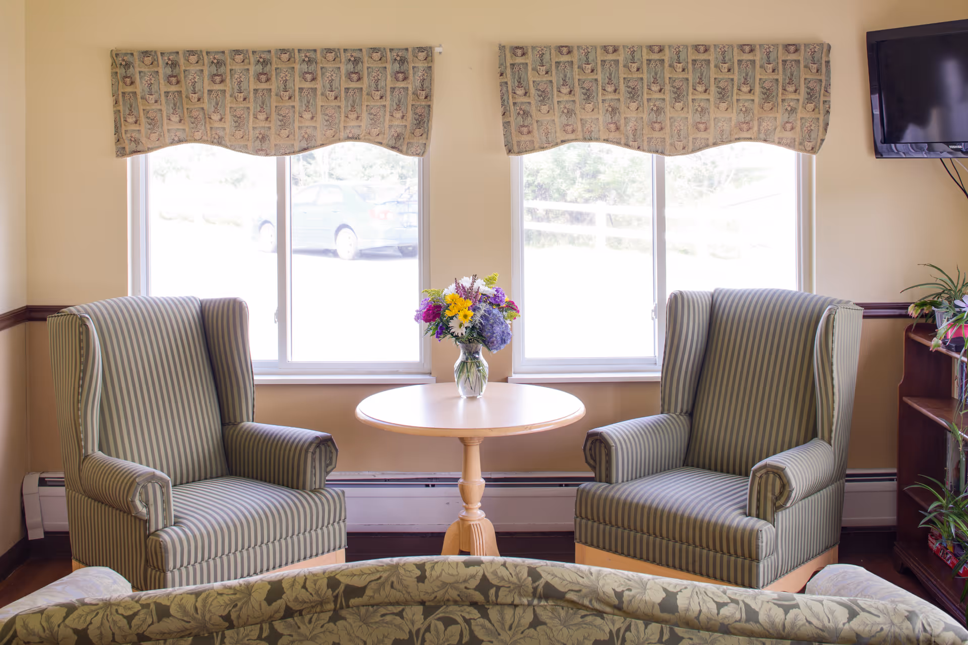 A cozy sitting area with two striped armchairs facing a small round wooden table with a vase of colorful flowers. Behind the chairs are two large windows with patterned valances, letting in natural light. A flat-screen TV is mounted on the wall to the right, and a bookshelf with plants is partially visible.