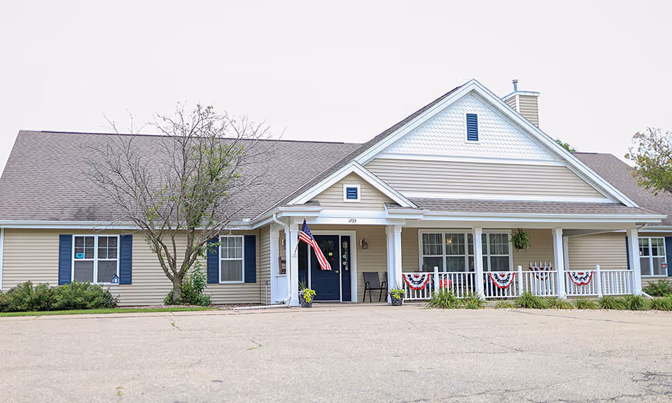 Front exterior view of a single-story senior living facility building with beige siding, blue shutters, a dark blue front door, an American flag, and a porch decorated with red, white, and blue bunting.