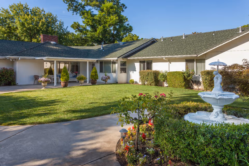 A sunny courtyard of an assisted living facility with a well-maintained lawn, potted plants, bushes, and a white decorative fountain featuring a statue holding an umbrella. The building surrounding the courtyard has white walls, green roof shingles, and several windows and doors.