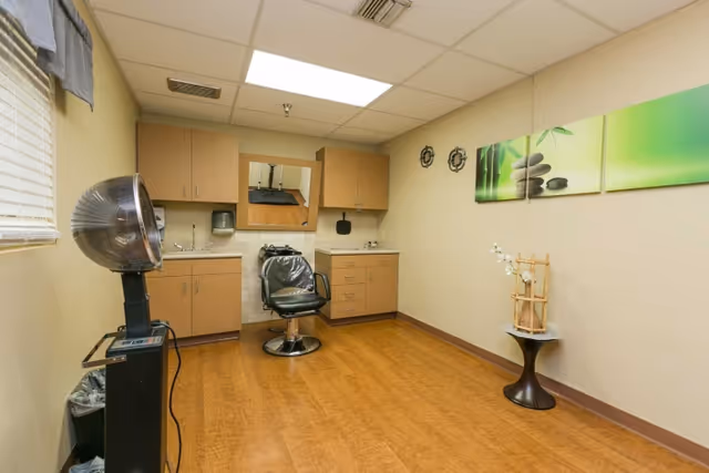 Interior view of a small salon or beauty room with a hair dryer chair, a black salon chair in front of a mirror, cabinets with sinks on either side, and a small table with decorative items. The walls are beige with a green-themed artwork and two small decorative wall hangings.