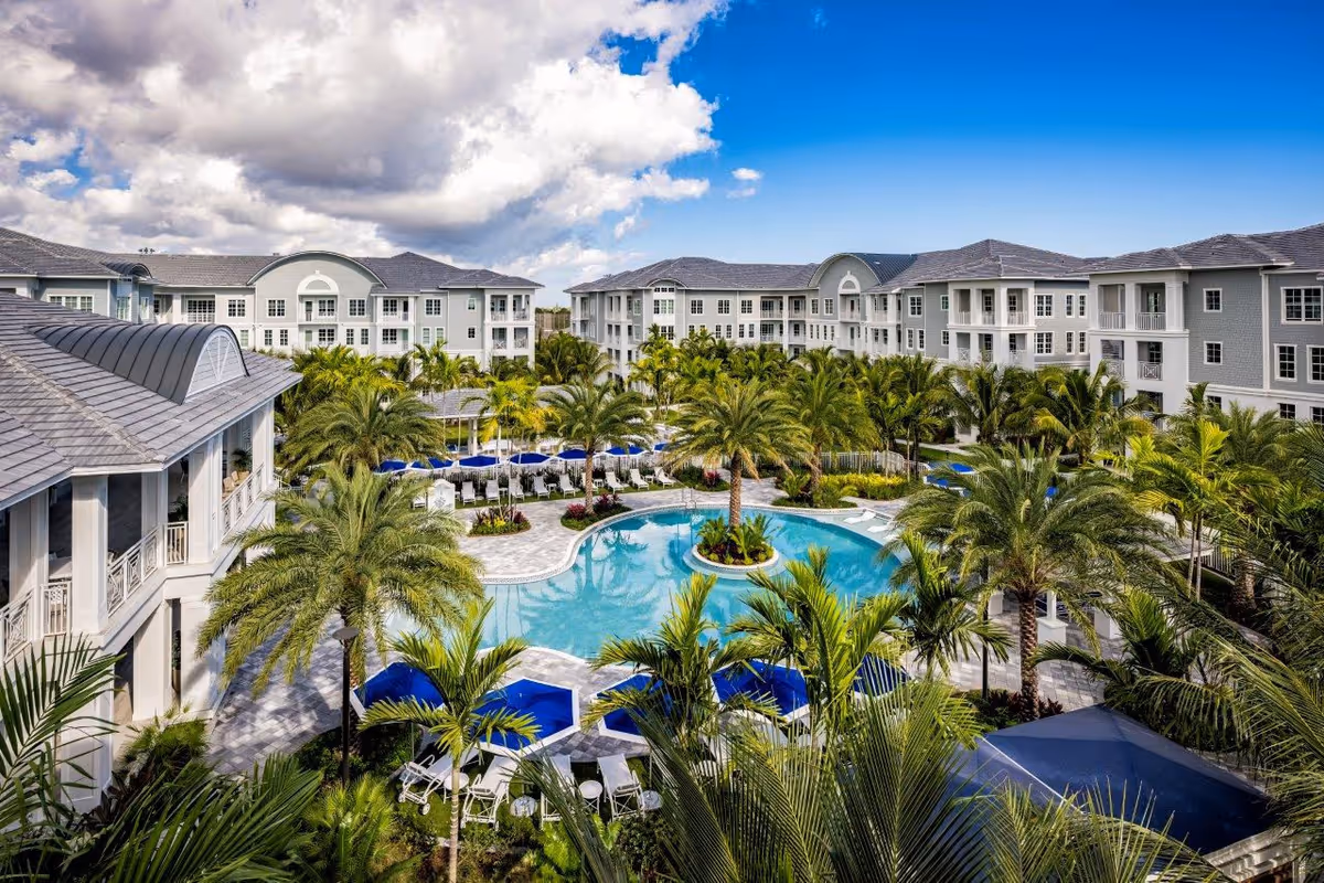 A large outdoor swimming pool surrounded by palm trees and lounge chairs with blue umbrellas, set within a senior living facility complex with multiple three-story buildings under a partly cloudy blue sky.