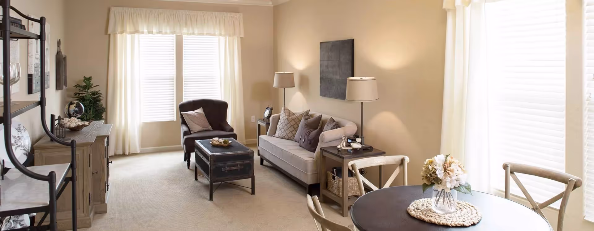 Bright, neutrally furnished living room and dining nook with a sofa, armchair, coffee table, side tables with lamps, and a round dining table by large windows.