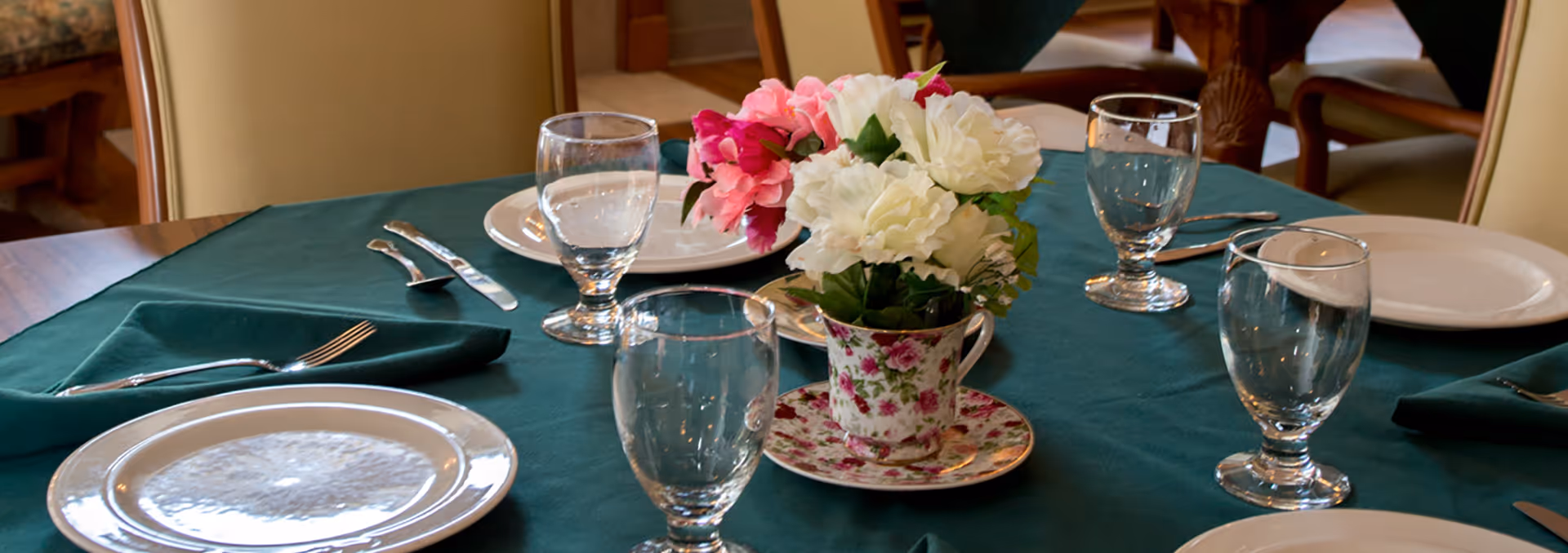A dining table set with plates, water glasses, silverware and a floral teacup centerpiece on a green tablecloth.