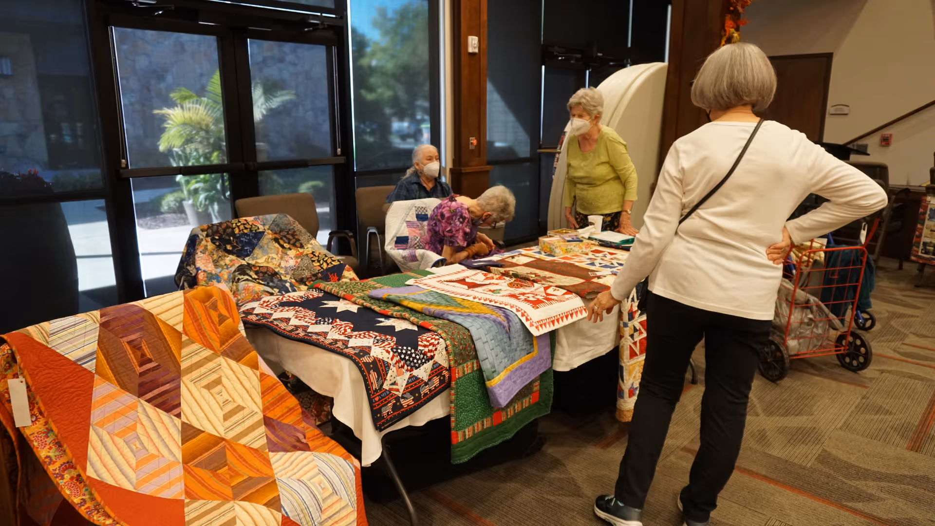 Several elderly women gathered around a table displaying colorful handmade quilts inside a well-lit room with large windows and carpeted floor. One woman is standing and looking at the quilts, while two others are seated behind the table, and another woman is standing behind them.