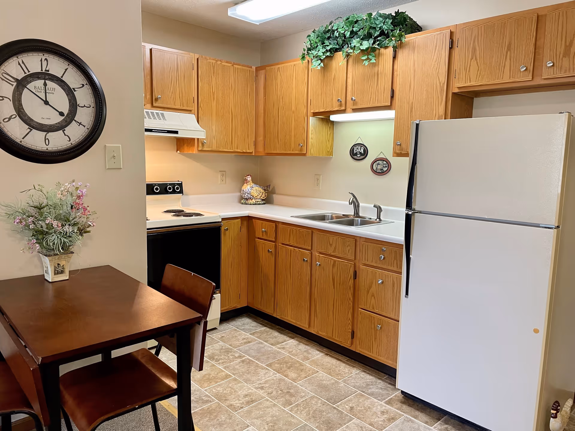 A small kitchen area with wooden cabinets, a white refrigerator, a stove with an overhead vent, a double sink, and a small dining table with two chairs. There is a wall clock above the table and a decorative plant on the table. Some greenery is placed on top of the cabinets.