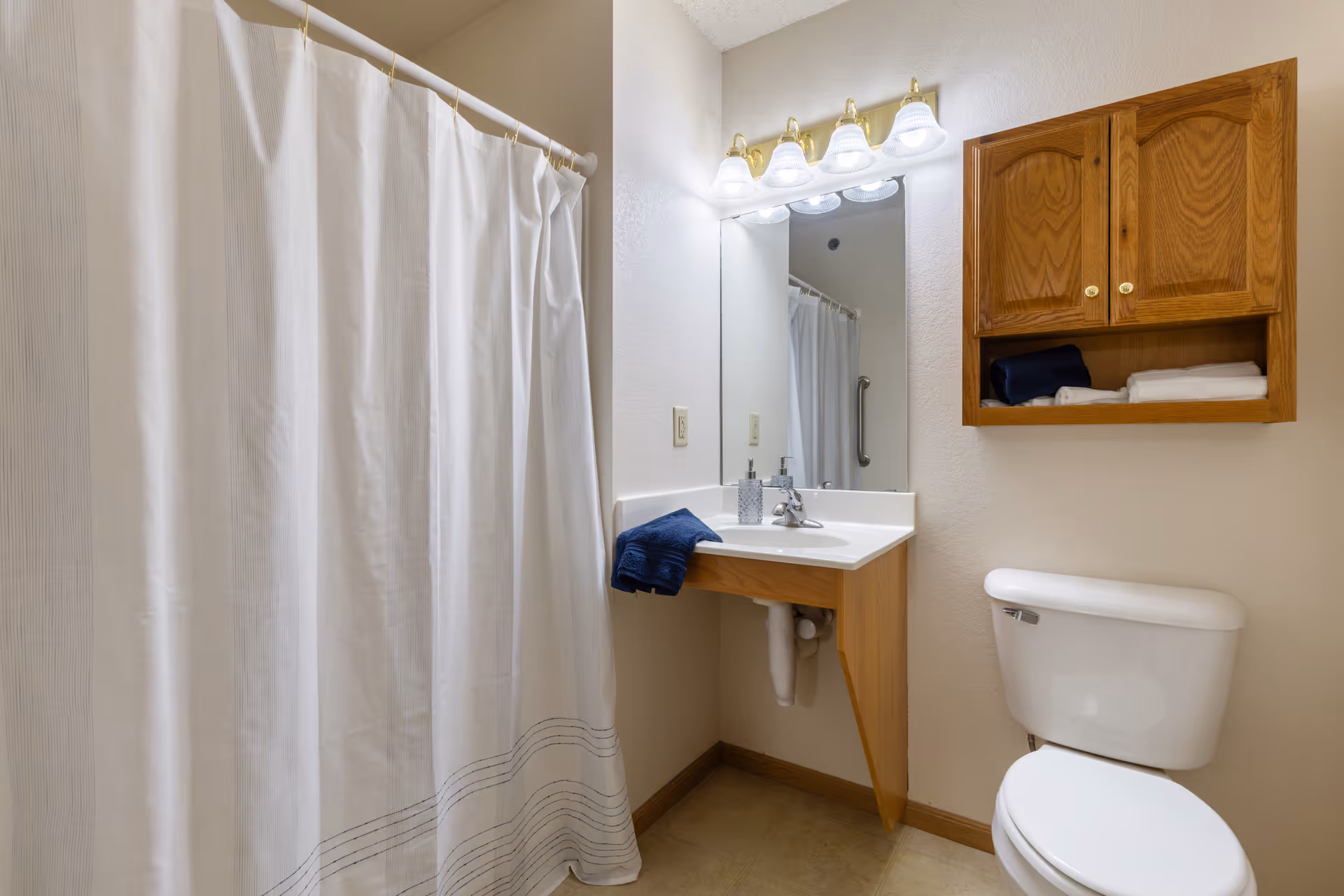 A clean bathroom featuring a white shower curtain, a white toilet, a wooden cabinet mounted on the wall with towels inside, a corner sink with a blue towel hanging on the side, a soap dispenser, and a large mirror with four light fixtures above it.