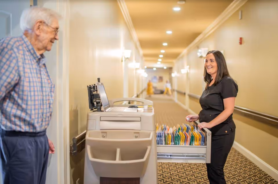 An elderly man in a plaid shirt stands in a hallway facing a smiling woman in black scrubs who is organizing files in a mobile cart with a laptop on top. The hallway is well-lit with beige walls and patterned carpet.