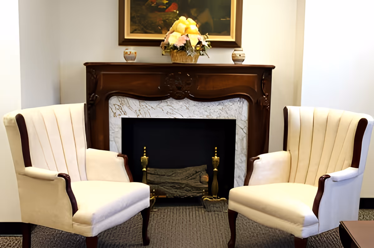 Two upholstered armchairs flank a decorative fireplace with a mantel, floral arrangement, and framed painting above.