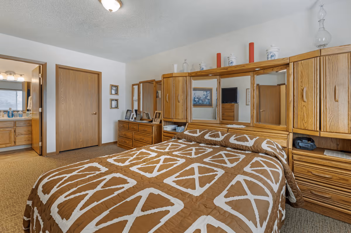 A bedroom in a senior living facility featuring a bed with a brown and white patterned bedspread. The room has wooden furniture including a dresser with a mirror, a headboard with cabinets and mirrors, and a nightstand. There are decorative items on top of the headboard and framed pictures on the walls. A door leads to an adjoining bathroom with a sink and mirror visible.
