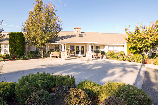 Front exterior view of Brookdale Torbett facility with a driveway, benches near the entrance, trees, and shrubs under a clear blue sky.