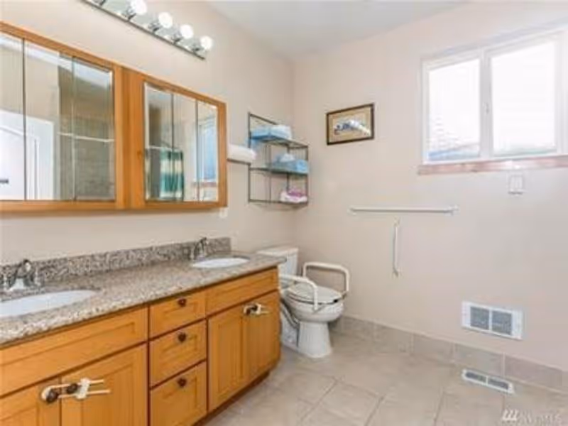 A spacious bathroom featuring a double sink vanity with granite countertop and wooden cabinets. There is a toilet with safety rails next to it, a wall-mounted shelf holding towels, a window letting in natural light, and a framed picture on the wall.